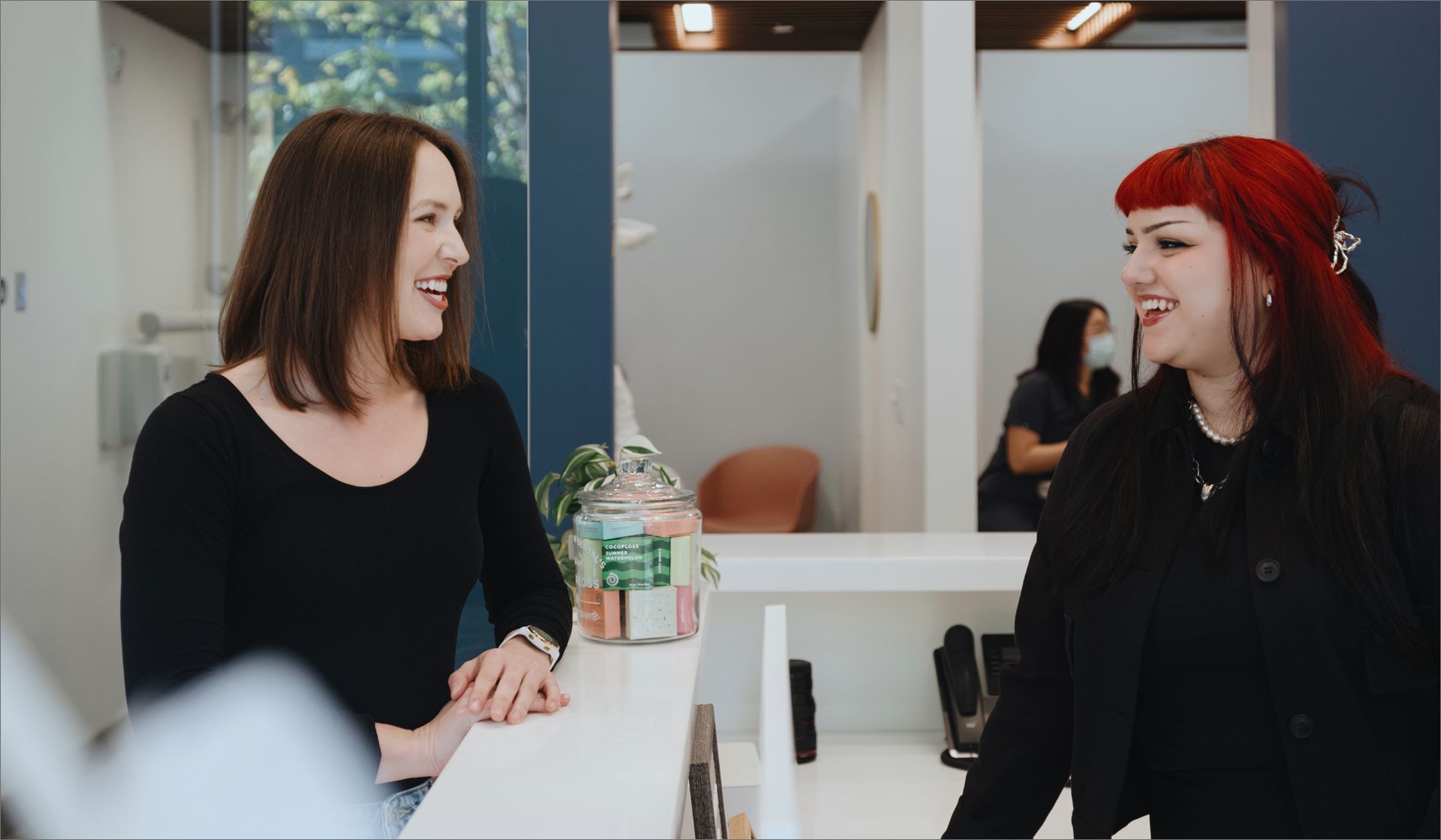 Two women smiling and talking across a white reception desk in a modern office setting.