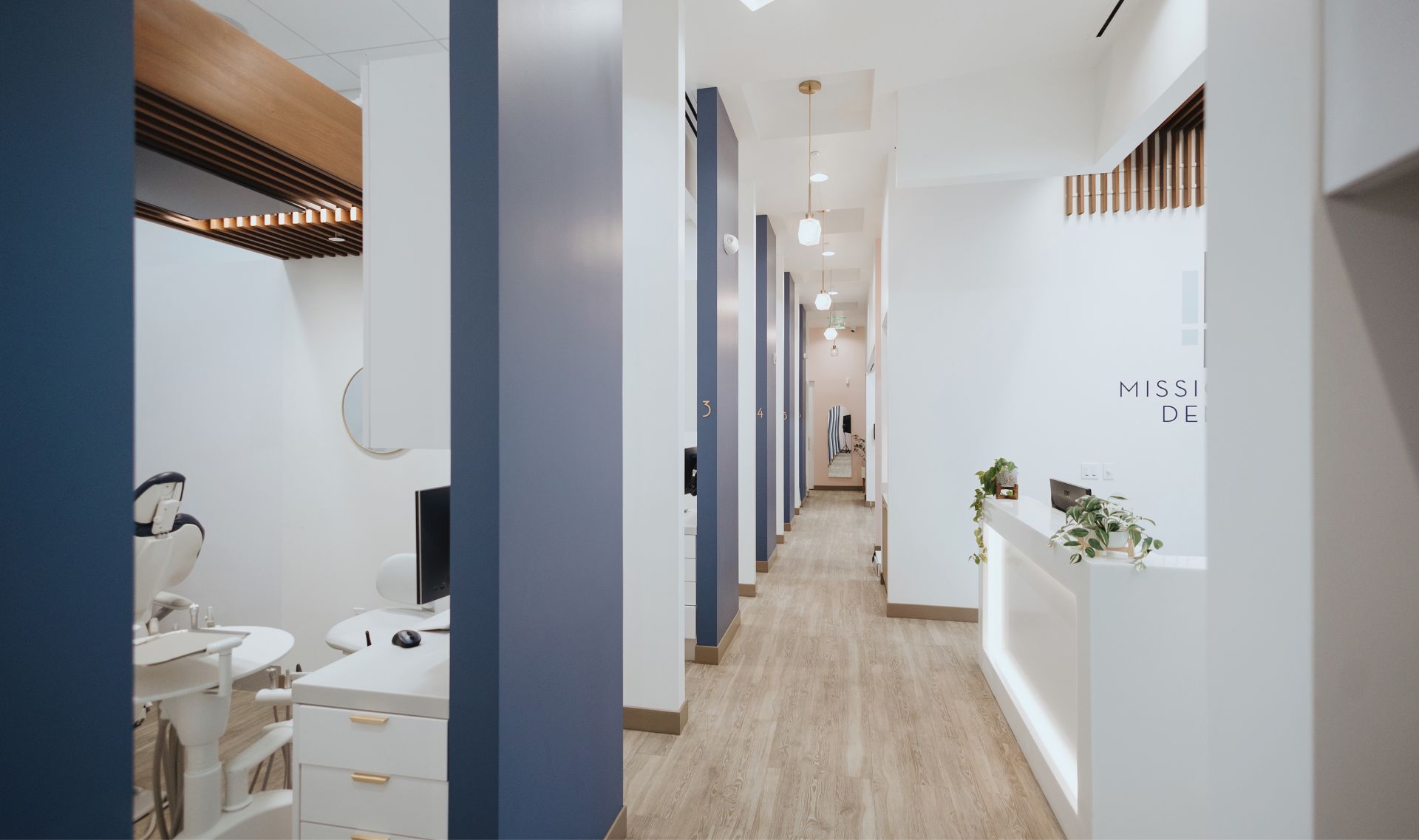 Modern dental office hallway with numbered blue and white booths, wooden floor, and white reception desk with plants.