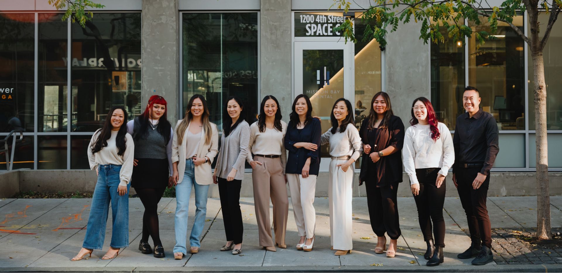 Group of ten smiling diverse adults standing in a row outside a building with glass walls and a sign reading '1200 4th Street SPACE'.