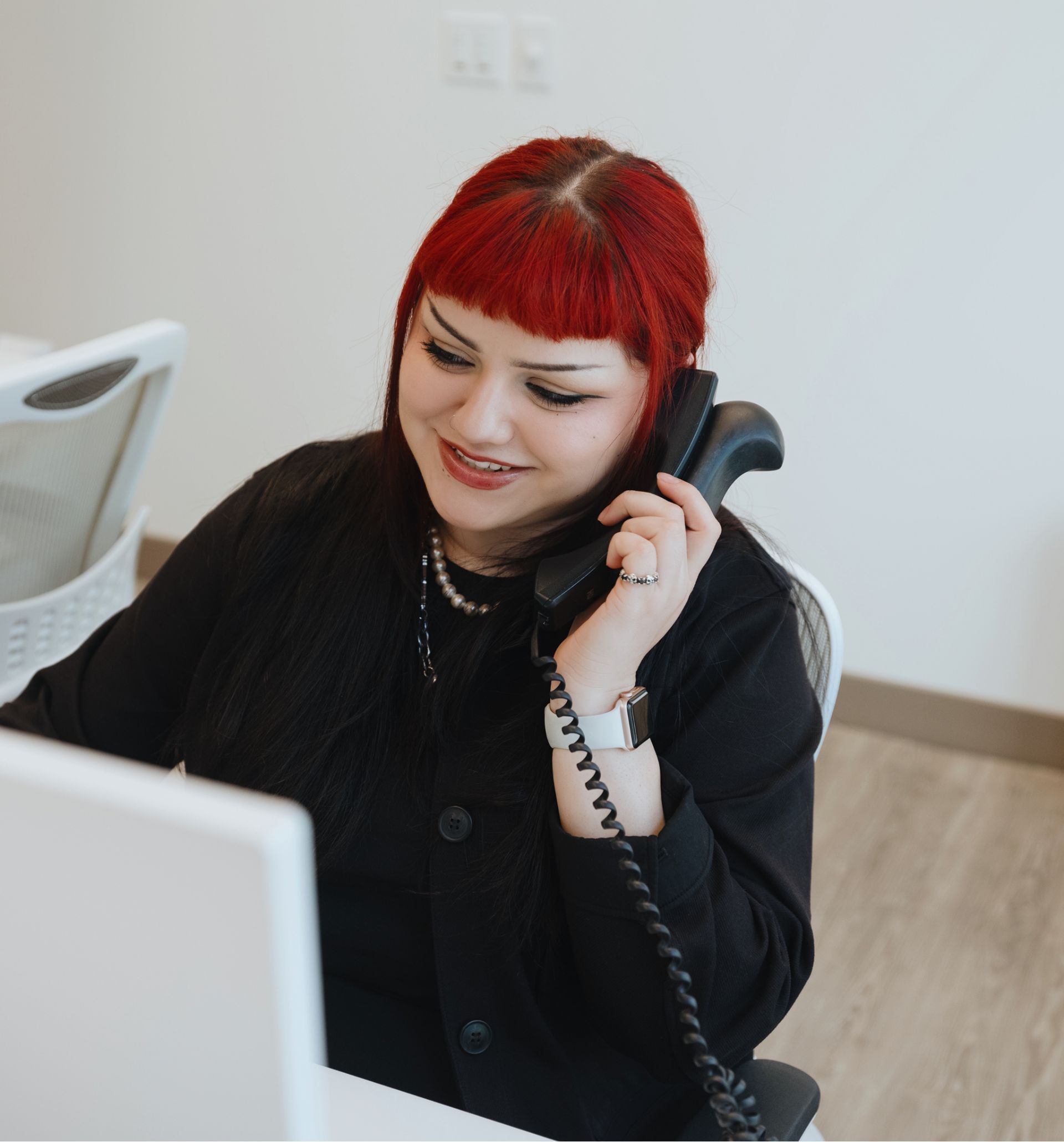 Smiling woman with red hair talking on a corded office phone while looking at a computer screen.