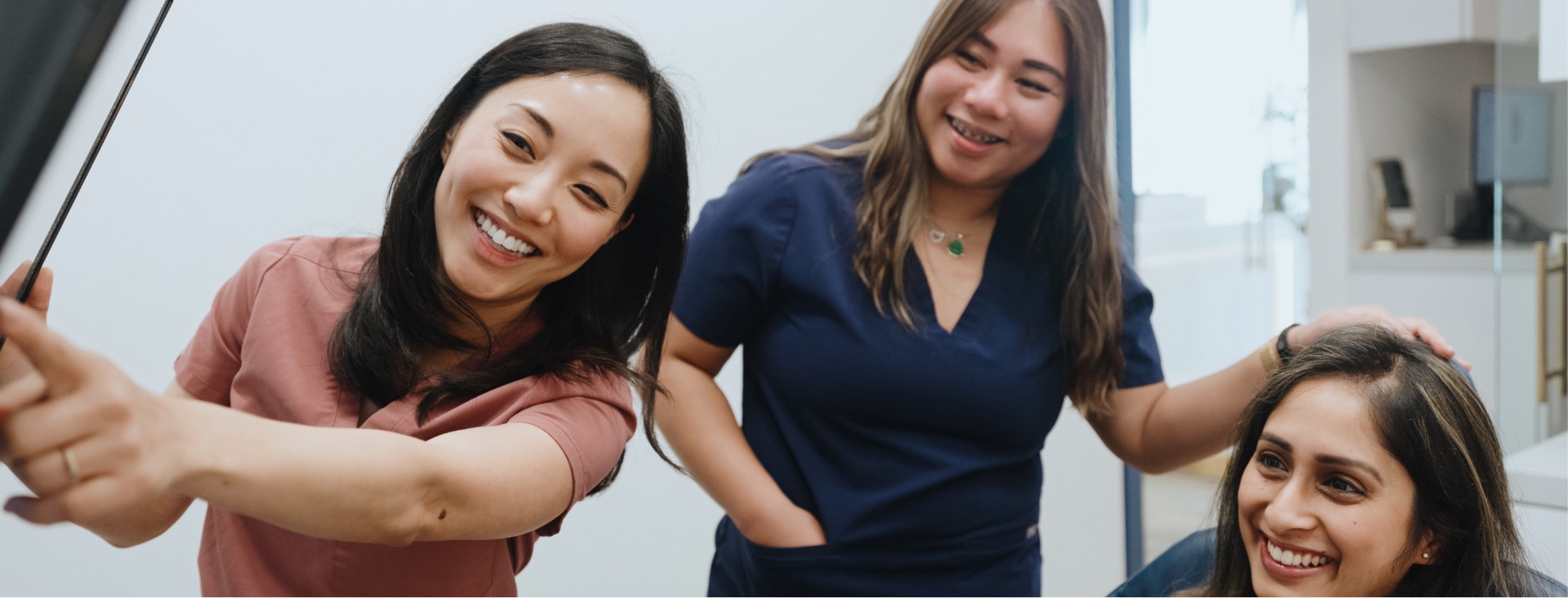 Three women smiling and interacting in a professional setting, one woman in scrubs touching another's head while the third takes a selfie.