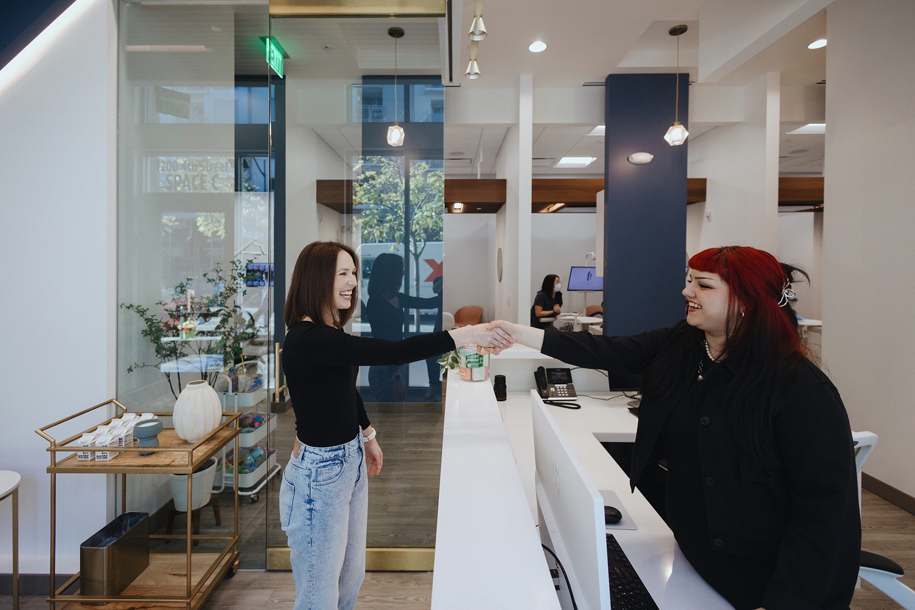 Two women smiling and shaking hands across a reception desk in a modern office or clinic.