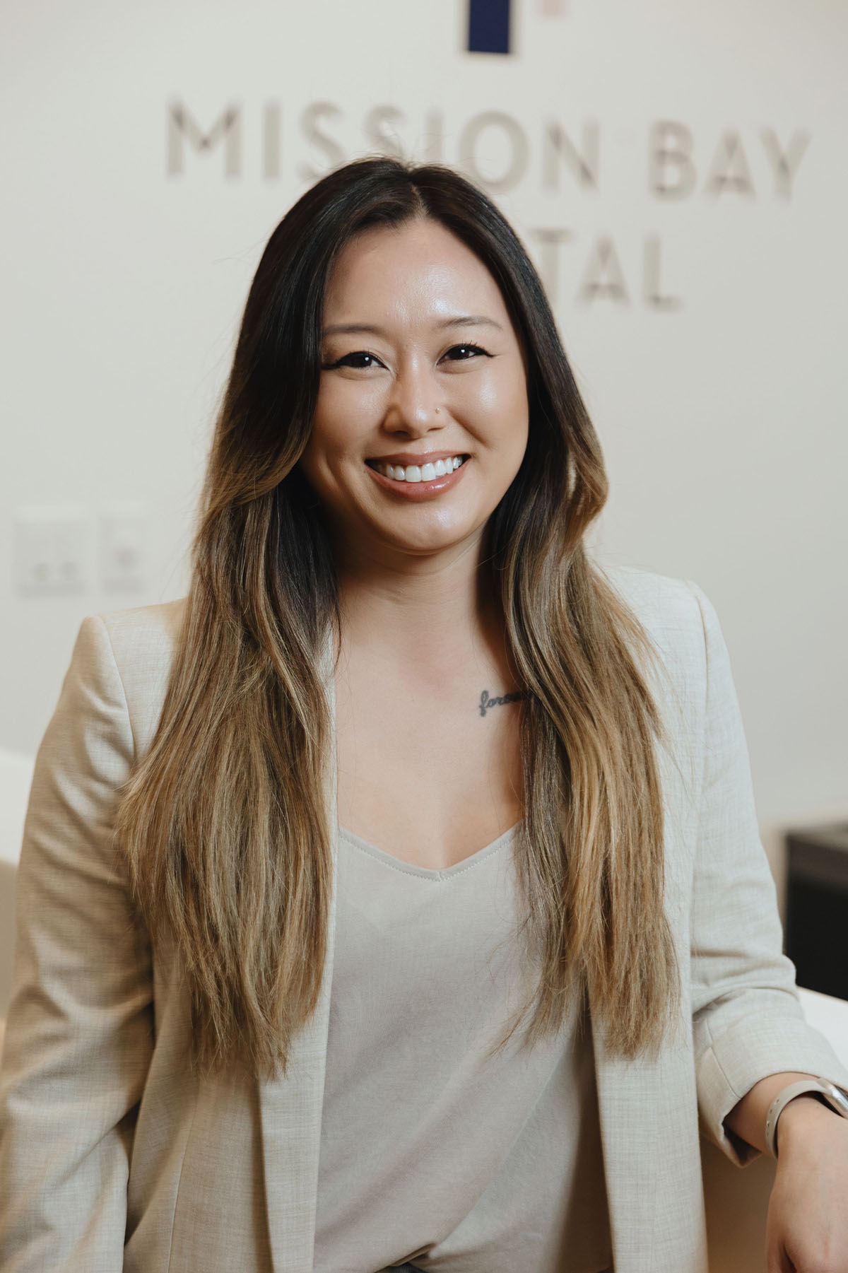 Smiling woman with long ombre hair wearing a beige blazer and tank top, sitting indoors with a Mission Bay Hospital sign in the background.