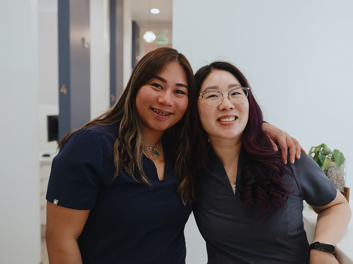 Two smiling women in dark scrubs embracing in a medical or office hallway.