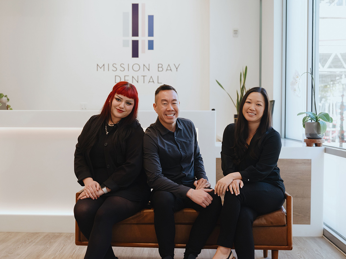 Three smiling dental professionals sitting on a brown bench in Mission Bay Dental reception area.