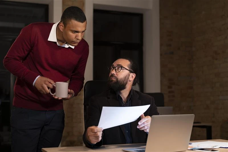 Homem de cabelo curto e camisa preta discutindo com colega de trabalho em um escritório moderno enquanto olha para uma folha de papel, com um laptop na mesa.