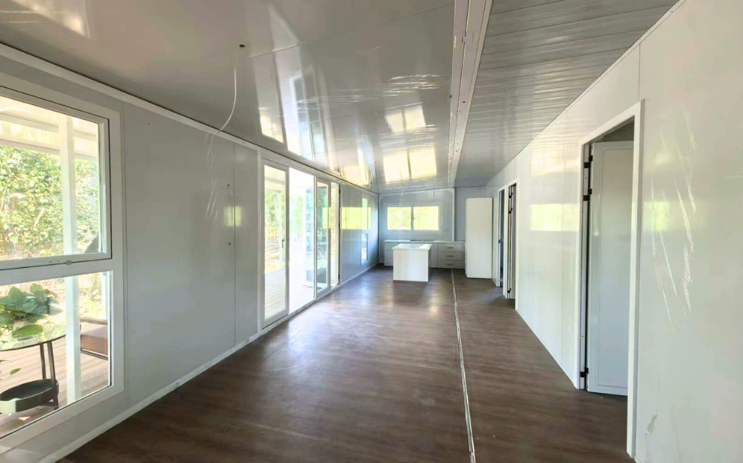 Interior of a modern cabin with large windows, white walls, wood flooring, and a small kitchen island at the far end.