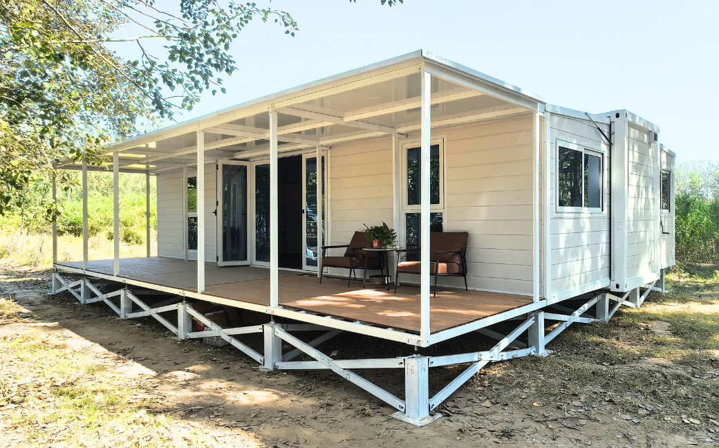 Modern white modular cabin elevated on metal supports with a covered porch featuring two brown chairs and a small table with a plant.