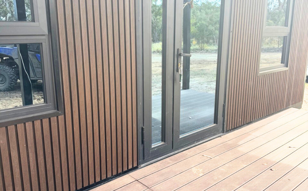 Close-up of a modern brown wooden exterior wall with glass double doors and windows, next to a wooden deck.