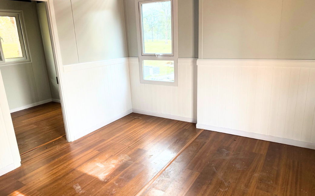 Empty room with wooden floor, white lower half walls, beige upper walls, and a window overlooking greenery.