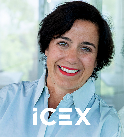 Mujer con cabello corto y oscuro, camisa azul clara y labios rojos, sonriente frente a fondo desenfocado con logo de ICEX.