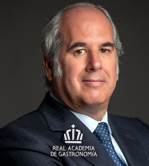 Retrato de un hombre calvo con cabello canoso, vestido con traje oscuro, camisa azul clara y corbata azul con puntos blancos, sobre fondo gris oscuro.