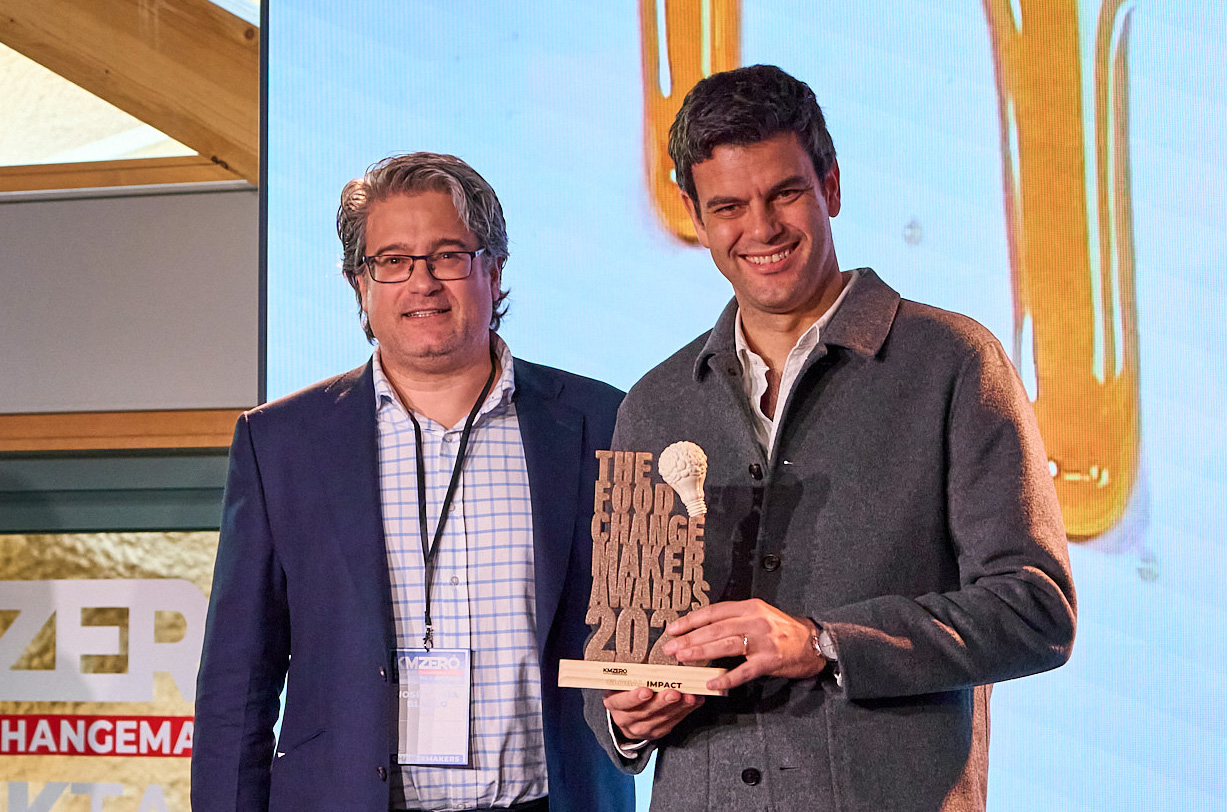 Dos hombres discutiendo documentos sentados en una cafetería, con personas conversando en el fondo.