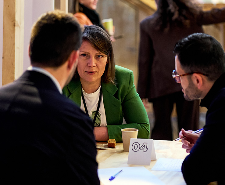 Dos hombres sentados en una mesa redonda discutiendo mientras uno usa gestos con las manos y frente a una tablet.