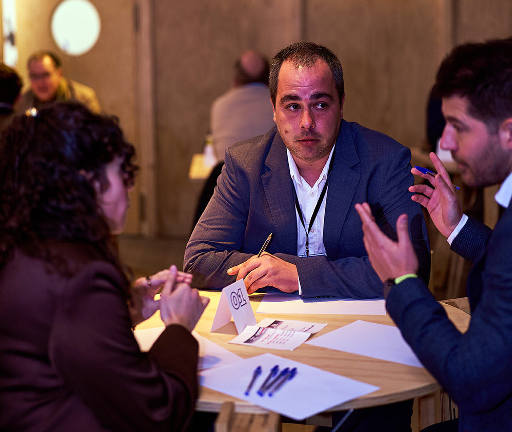 Dos hombres sentados en una mesa redonda discutiendo mientras uno usa gestos con las manos y frente a una tablet.