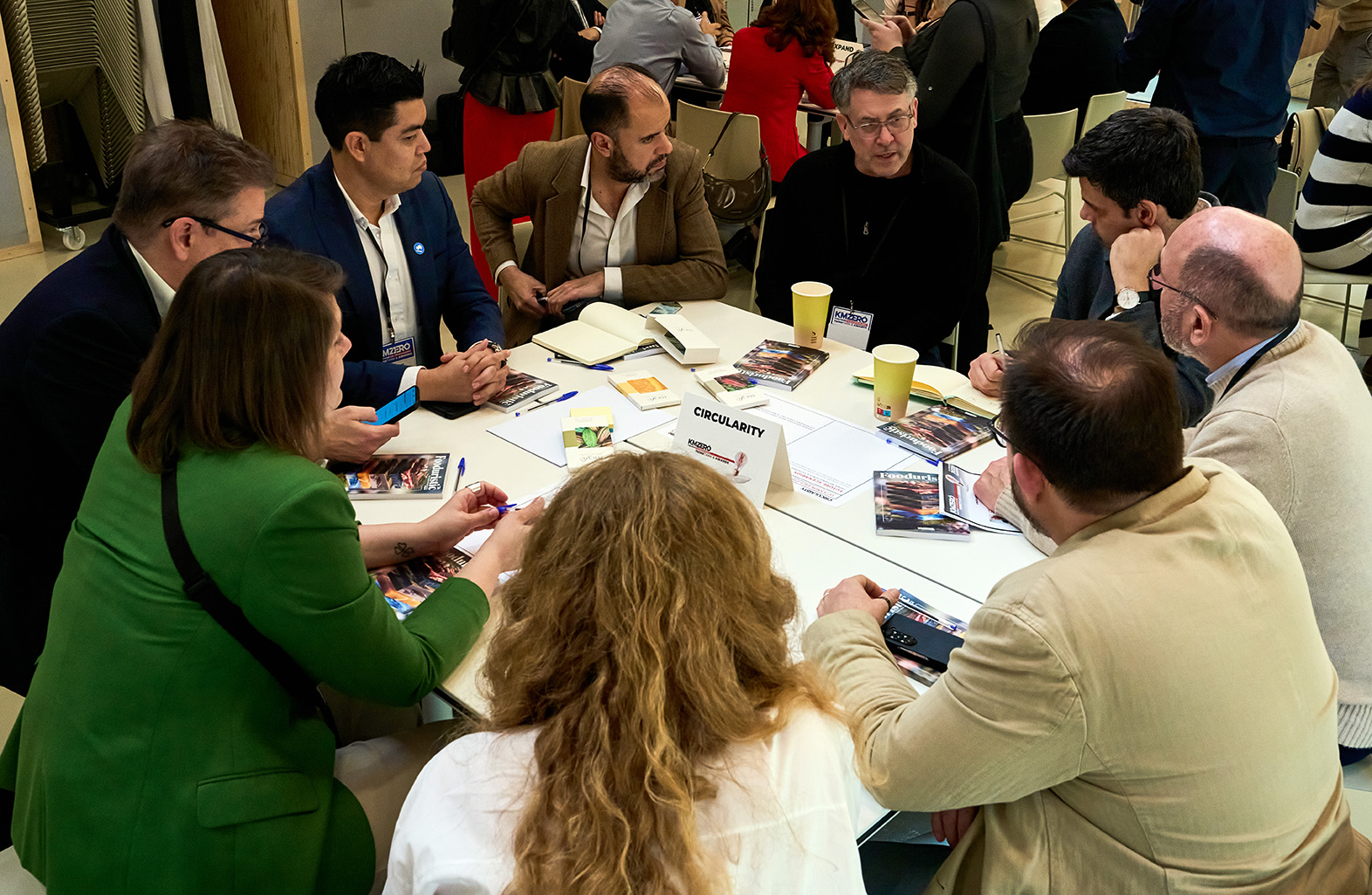 Dos hombres sentados en una mesa redonda discutiendo mientras uno usa gestos con las manos y frente a una tablet.