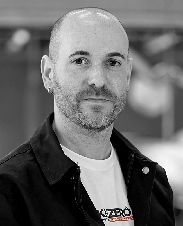 Hombre calvo con camisa negra de manga corta sonriendo con los brazos cruzados en un entorno industrial.