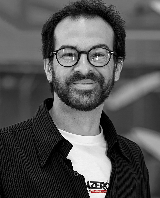 Hombre con gafas y barba ligera sonriendo, con camisa de rayas oscuras y fondo desenfocado.