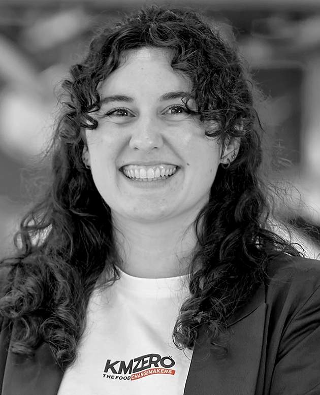 Mujer sonriente con cabello rizado y camiseta negra con el texto KMZERO, brazos cruzados.
