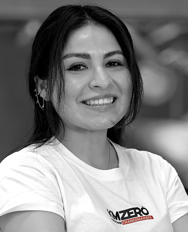 Mujer joven sonriendo con cabello suelto y ropa negra, con collar y aretes, en un entorno al aire libre desenfocado.
