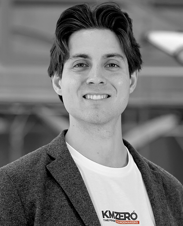 Hombre calvo con camisa negra de manga corta sonriendo con los brazos cruzados en un entorno industrial.