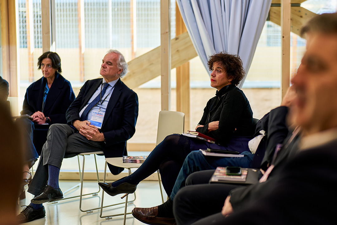 Dos hombres discutiendo documentos sentados en una cafetería, con personas conversando en el fondo.