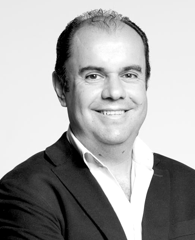 Hombre sonriendo con los brazos cruzados, vestido con chaqueta oscura y camisa blanca.