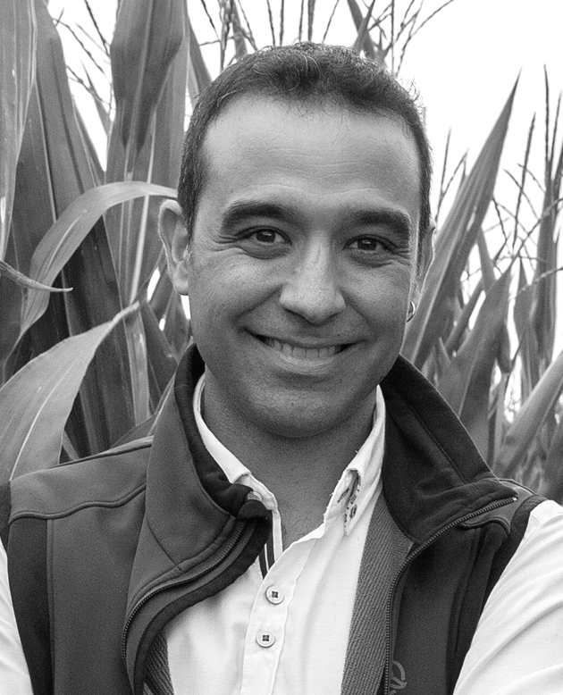 Hombre sonriendo con los brazos cruzados, vestido con chaqueta oscura y camisa blanca.