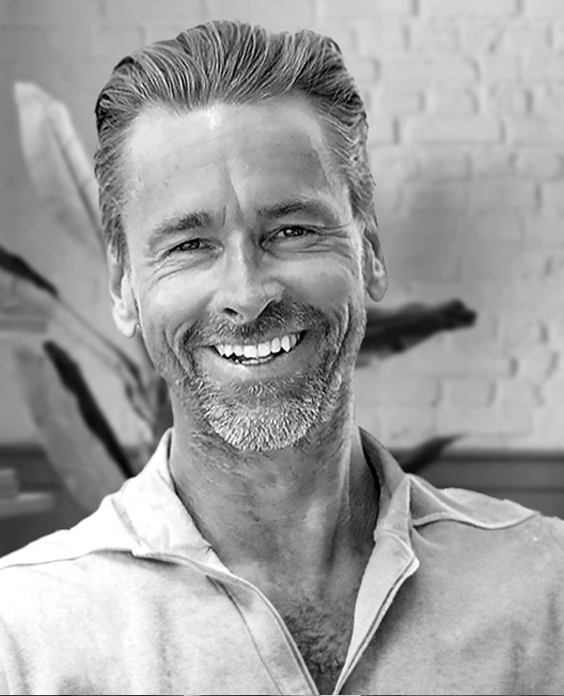 Hombre sonriendo con los brazos cruzados, vestido con chaqueta oscura y camisa blanca.
