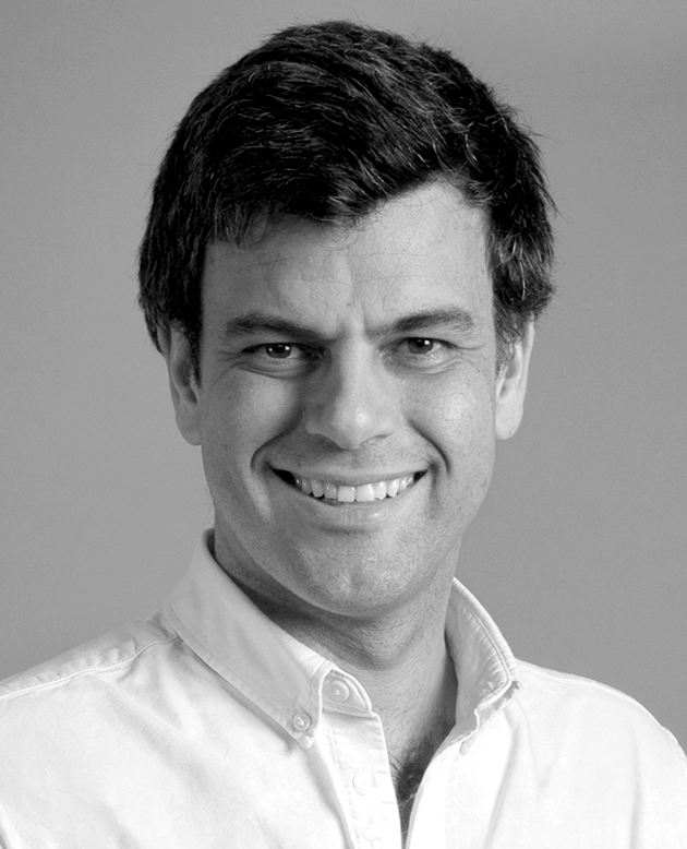 Hombre sonriendo con los brazos cruzados, vestido con chaqueta oscura y camisa blanca.