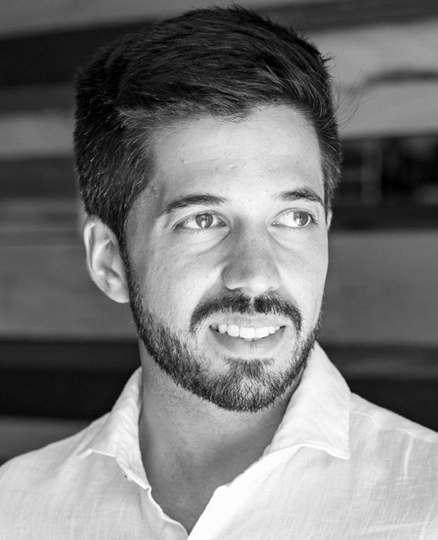 Hombre sonriendo con los brazos cruzados, vestido con chaqueta oscura y camisa blanca.