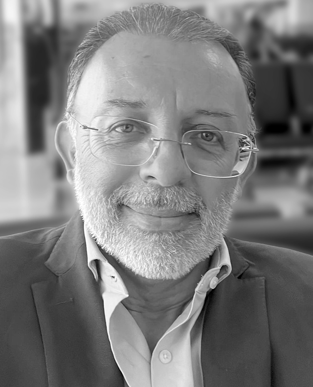 Hombre sonriendo con los brazos cruzados, vestido con chaqueta oscura y camisa blanca.