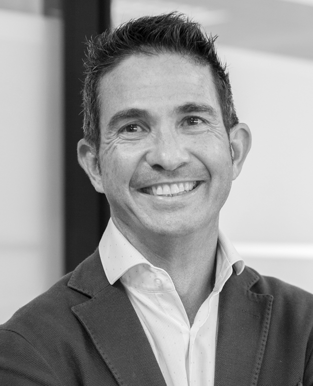 Hombre sonriendo con los brazos cruzados, vestido con chaqueta oscura y camisa blanca.