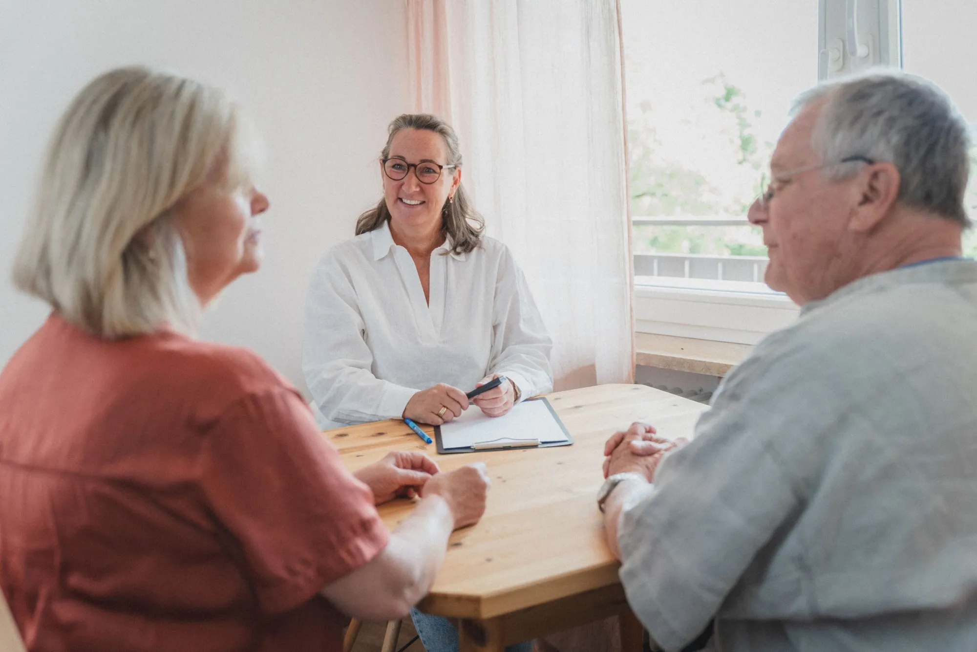 Beraterin spricht mit einem älteren Paar an einem Holztisch in einem hellen Raum.