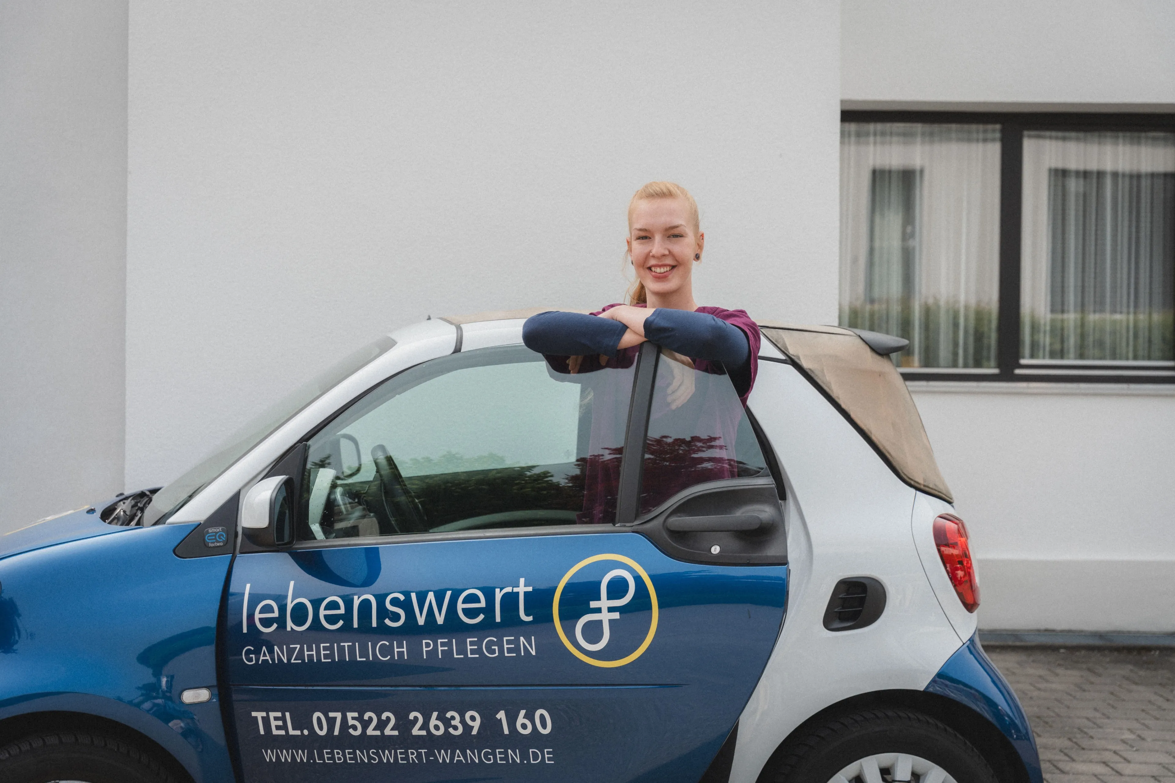 Pflegekraft lehnt lächelnd am geöffneten Autotür eines blauen und weißen Fahrzeugs mit der Aufschrift lebenswert ganzheitlich pflegen.