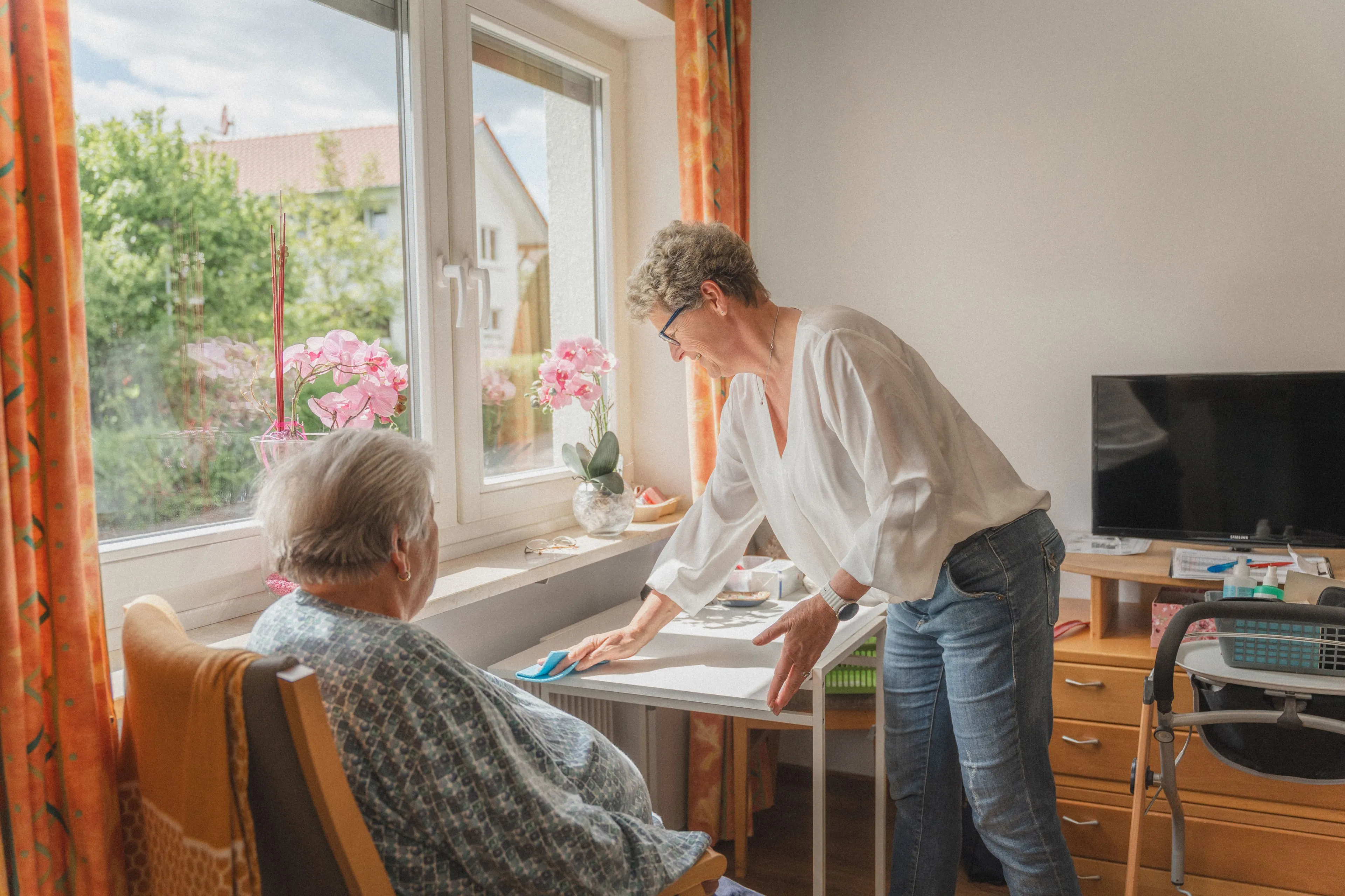 Eine Frau putzt einen kleinen Tisch in einem Zimmer, während eine ältere Frau auf einem Stuhl sitzt und zuschaut.