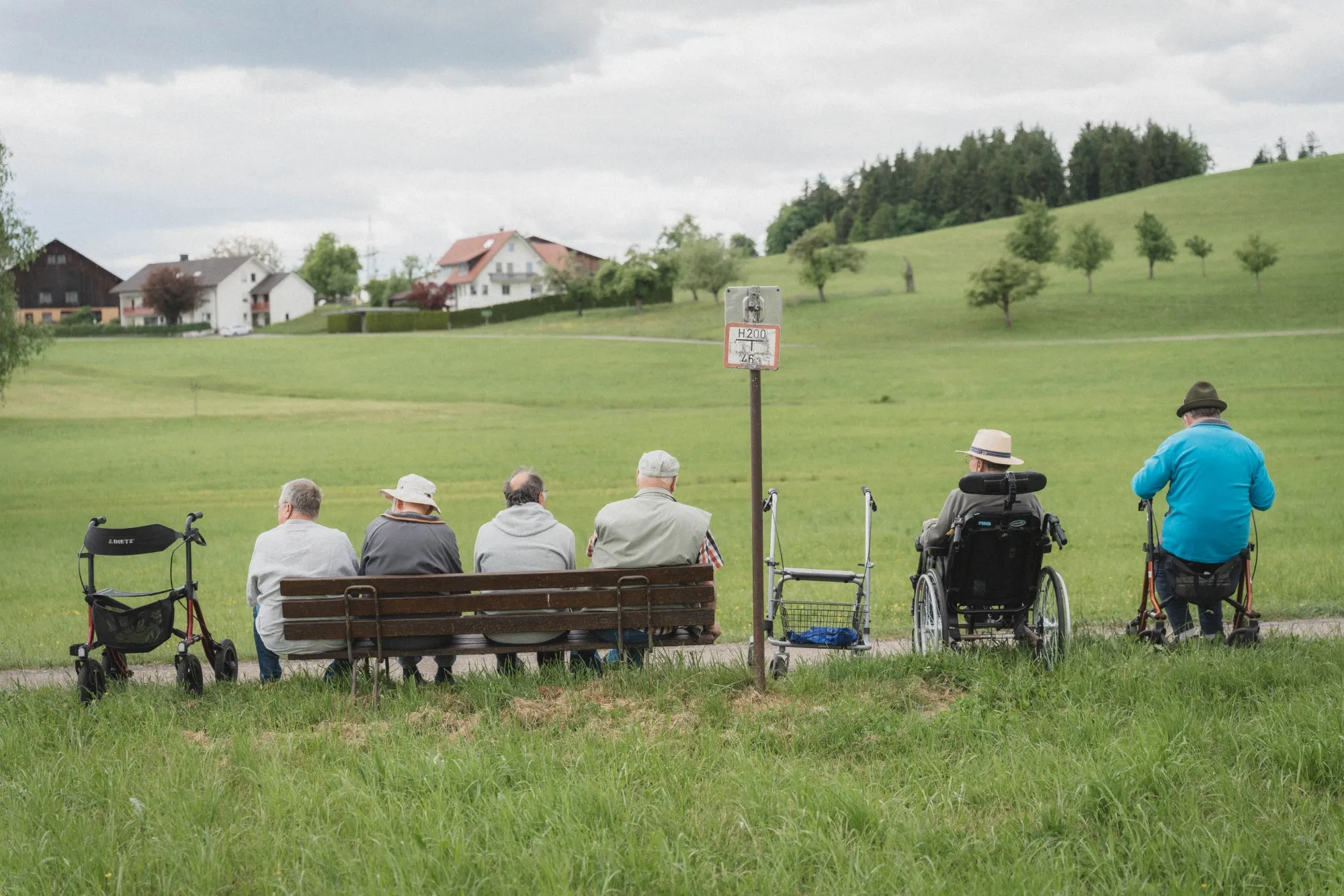 Mehrere ältere Menschen sitzen auf einer Bank und Rollstühlen vor einer grünen Wiese mit Häusern im Hintergrund.