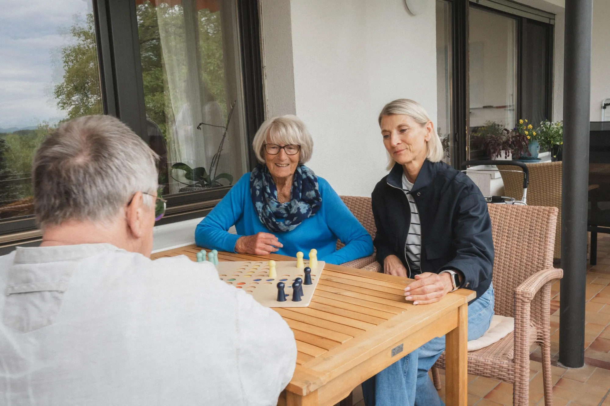 Drei ältere Erwachsene spielen ein Brettspiel zusammen an einem Holztisch auf einer Terrasse.