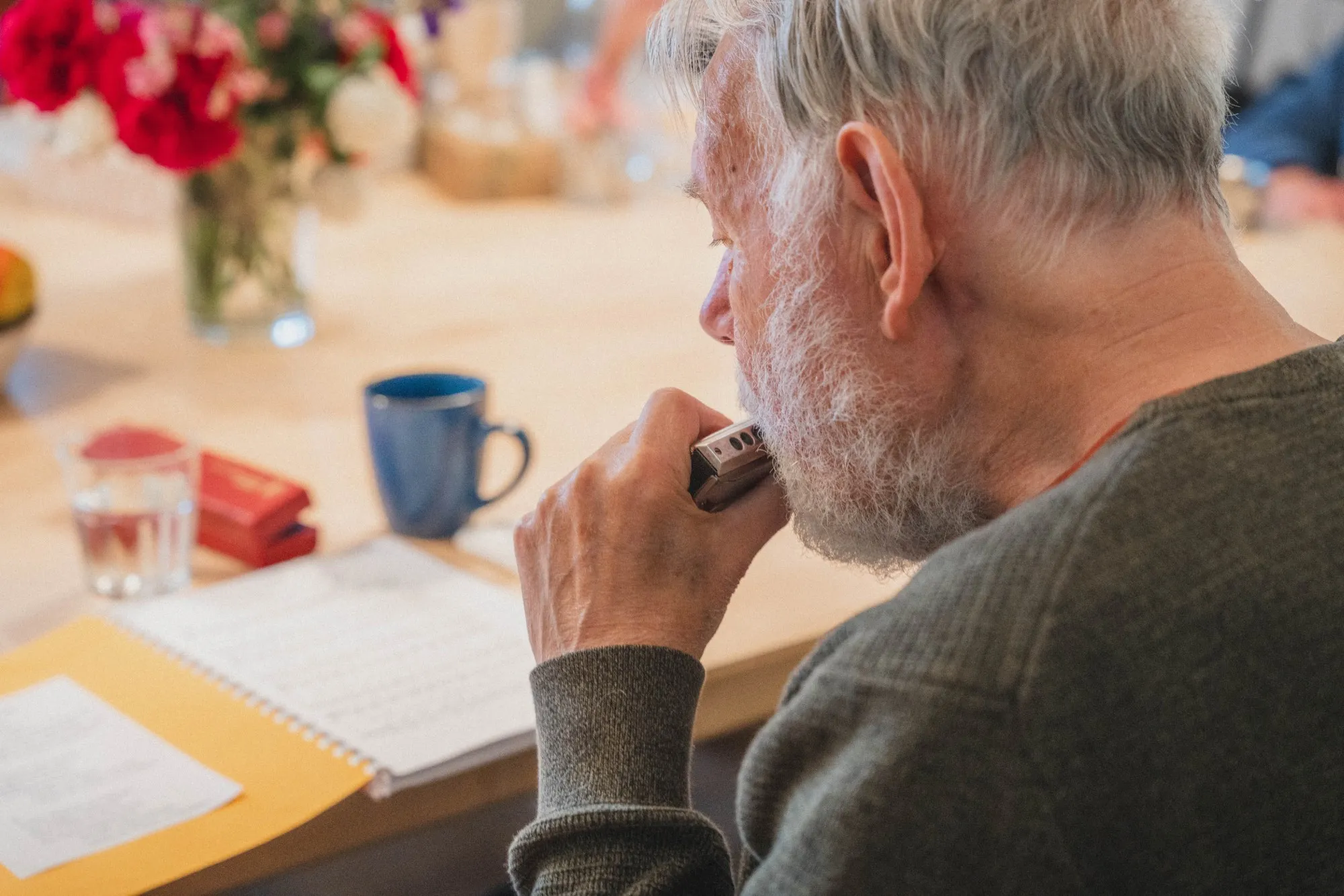 Älterer Mann spielt Mundharmonika an einem Tisch mit Noten, Tasse und Glas Wasser.