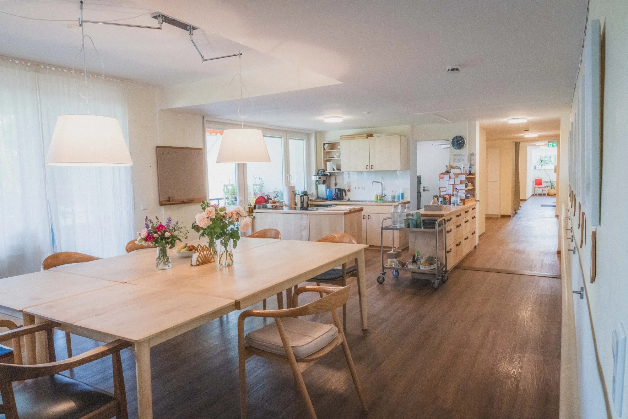 Heller Gemeinschaftsbereich mit großem Esstisch, Holzstühlen, Blumenvasen und moderner Küchenzeile im Hintergrund.