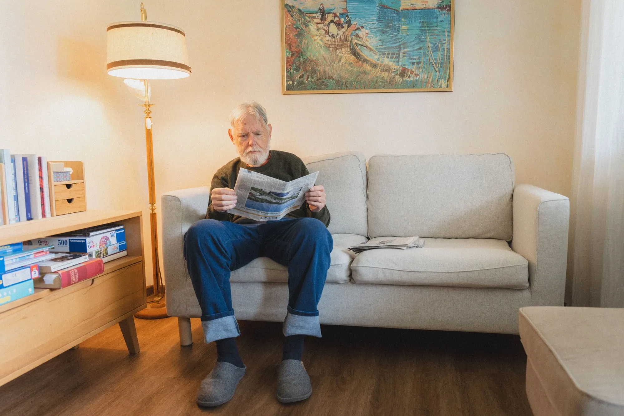Älterer Mann sitzt auf einem grauen Sofa in gemütlichem Wohnzimmer und liest Zeitung.