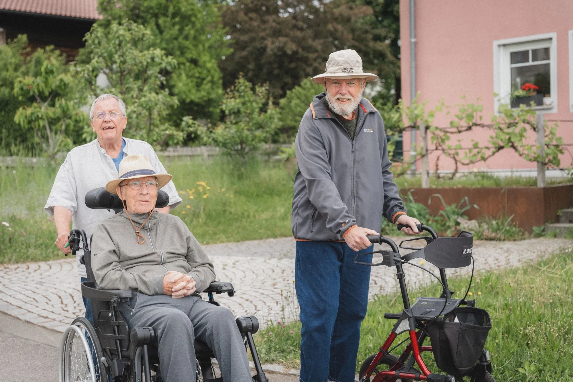 Drei Senioren im Freien, einer im Rollstuhl, einer mit Rollator, umgeben von Grün und einem rosa Haus im Hintergrund.