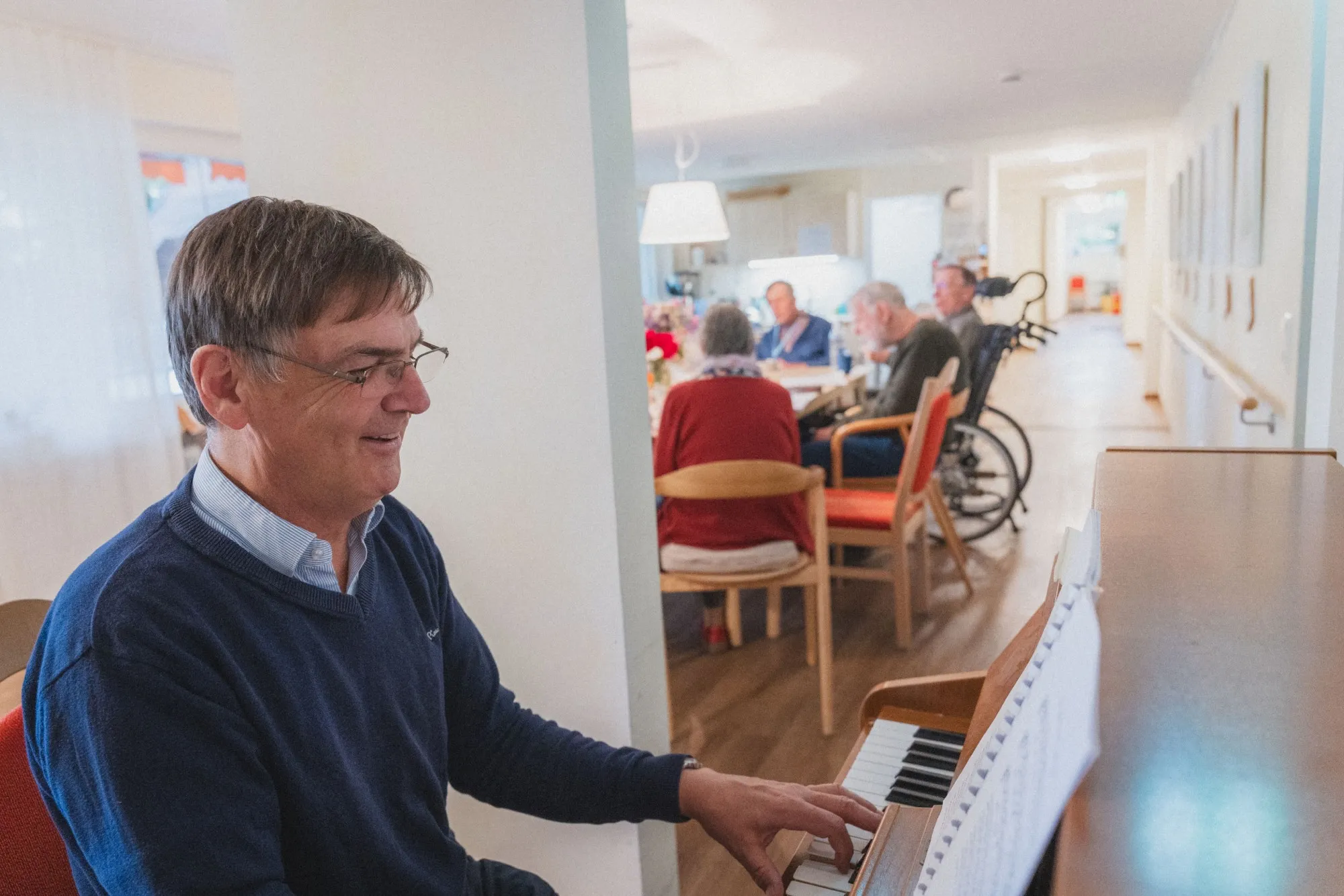 Ein älterer Mann spielt am Klavier, während im Hintergrund mehrere Senioren an einem Tisch sitzen.