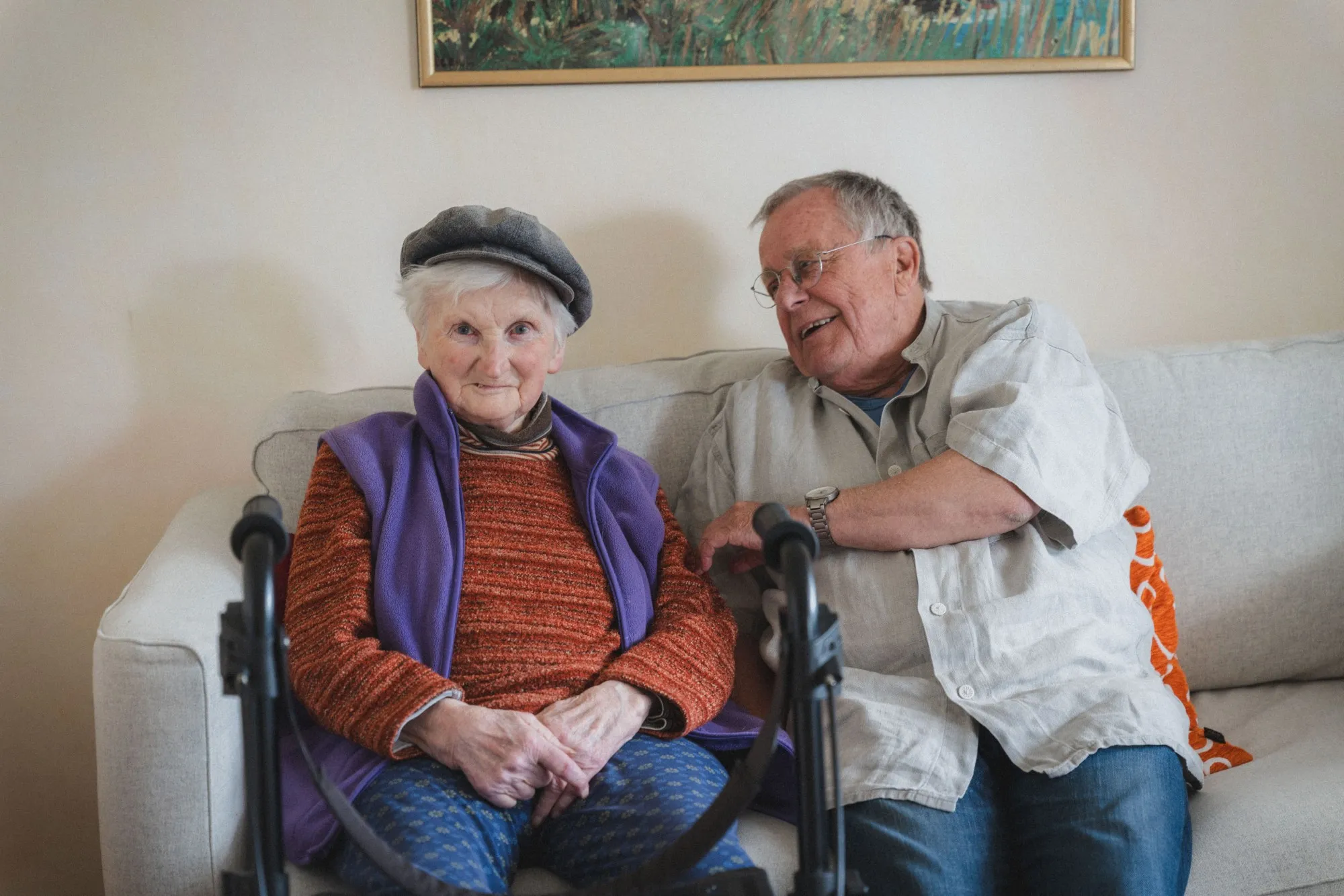 Ältere Frau mit Mütze und lila Weste sitzt auf einem Sofa neben einem älteren Mann mit Brille, der sie anlächelt.