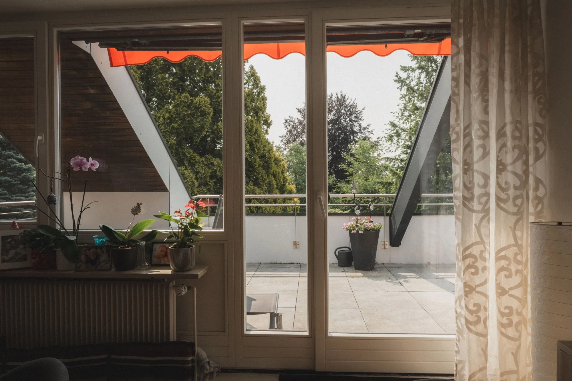 Blick von einem Innenraum mit Blumen auf einem Fensterbrett auf eine Terrasse mit Pflanzen und Bäumen im Hintergrund.