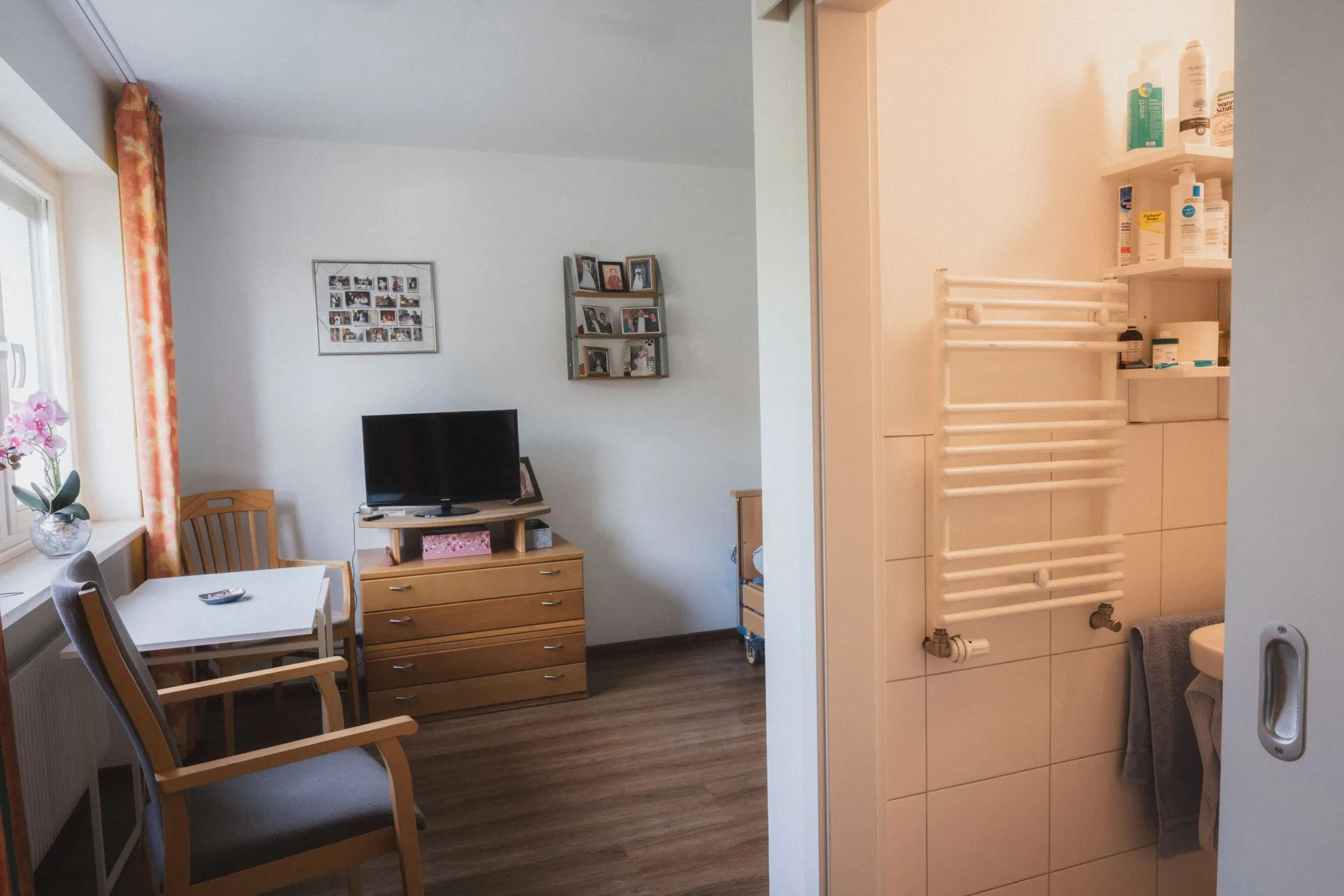 Gemütlicher Raum mit zwei Stühlen, einem kleinen Tisch, einer Kommode mit Fernseher und einem Blick in ein angrenzendes Bad mit Handtuchwärmer und Regal.