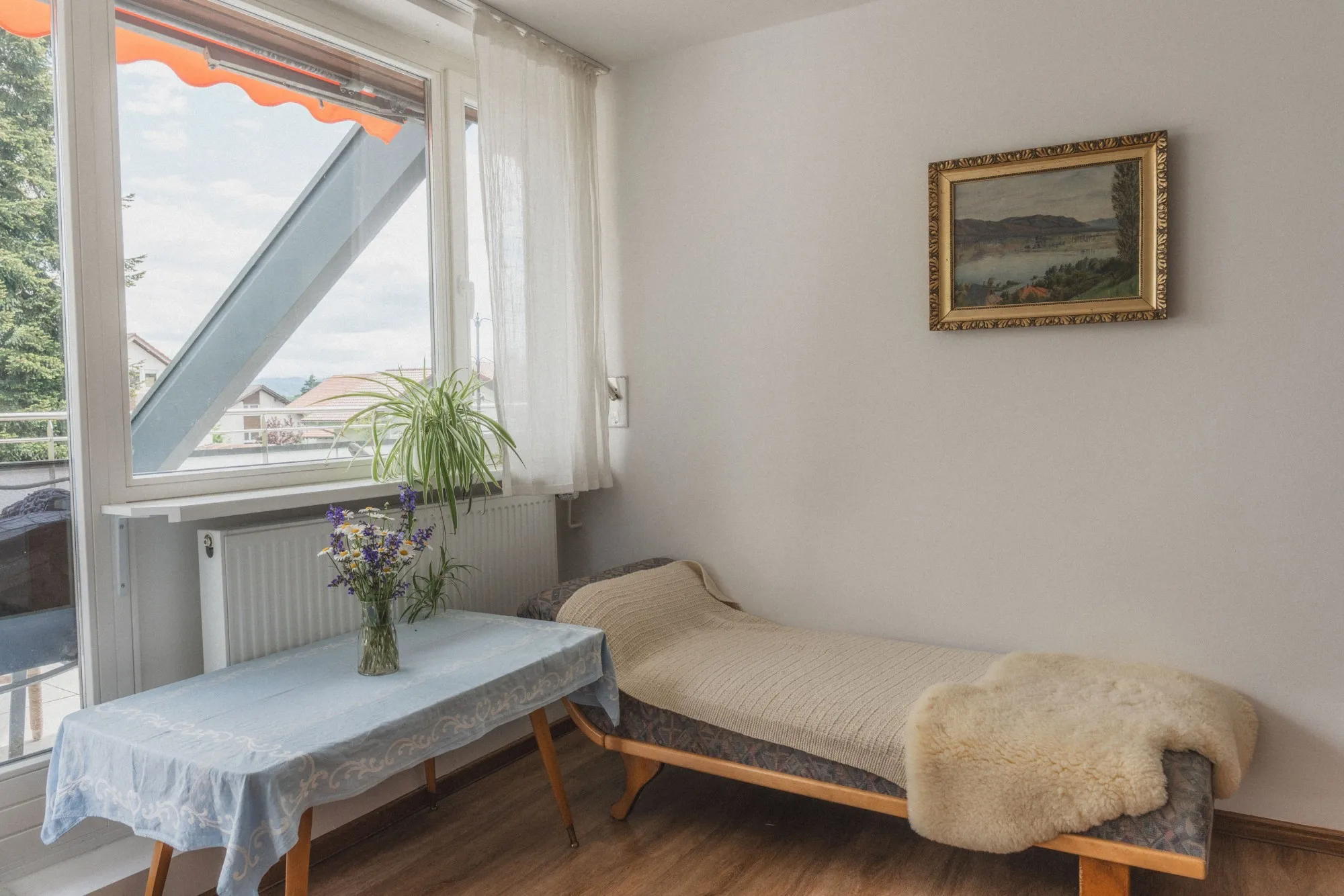 Heller Raum mit einem Holzsofa, das mit einer Decke und einem Fell bedeckt ist, einem Tisch mit einer blauen Tischdecke und Vase mit Blumen neben einem großen Fenster.