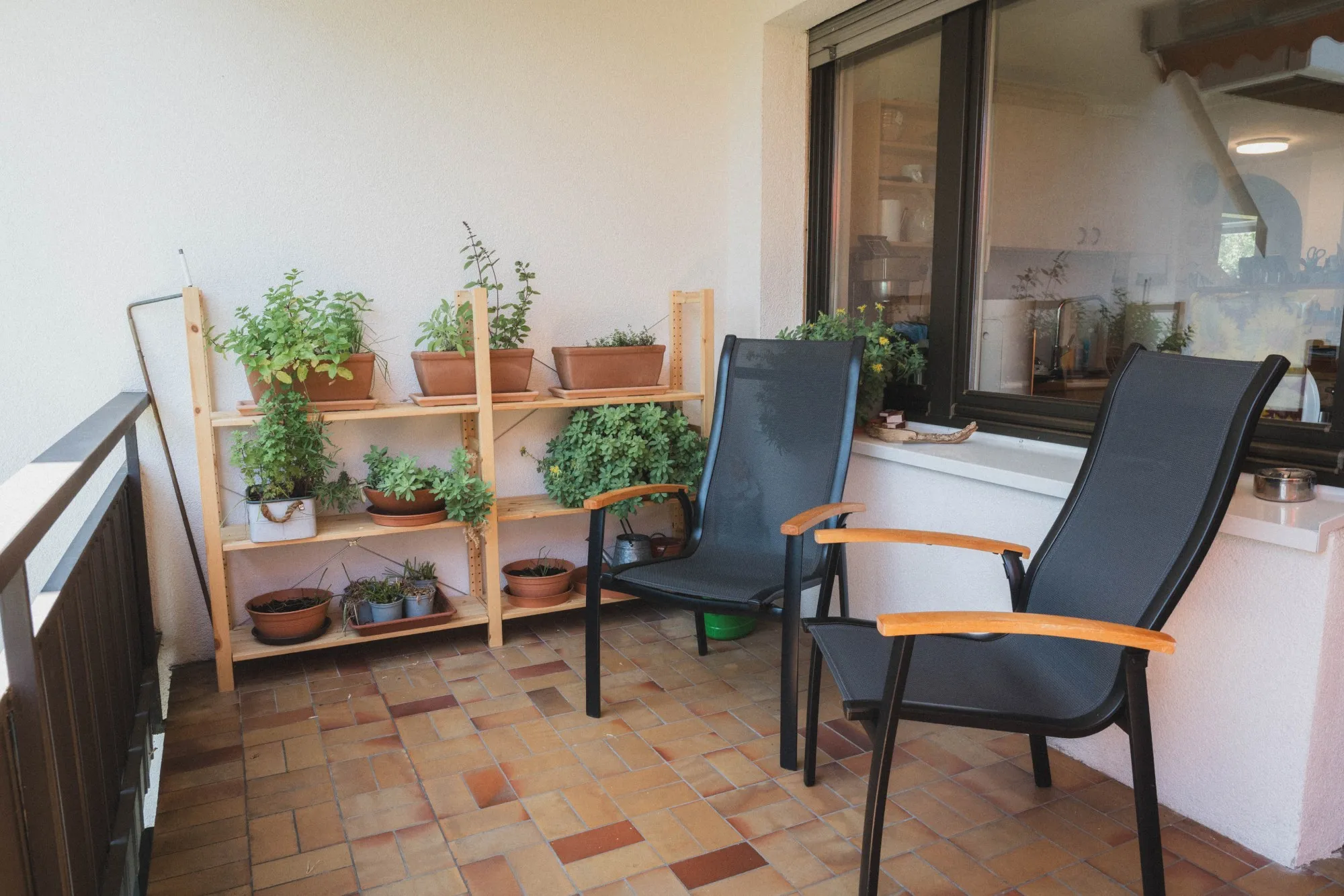 Balkon mit zwei schwarzen Stühlen mit Holzarmlehnen neben einem Regal mit verschiedenen Pflanzen in Blumentöpfen.