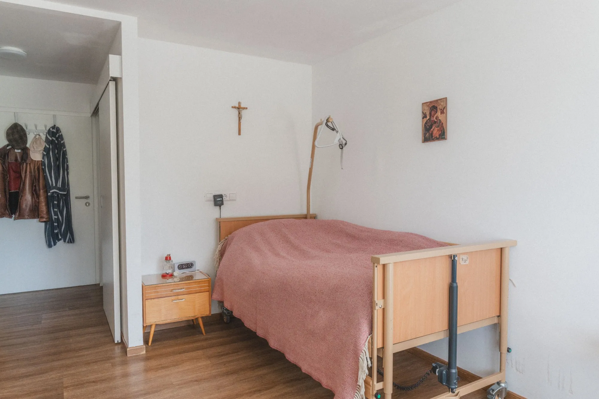 Ein einfach eingerichtetes Zimmer mit einem Pflegebett, einem Nachttisch, einem roten Deckenbezug, einem Kreuz und einem religiösen Bild an der weißen Wand.