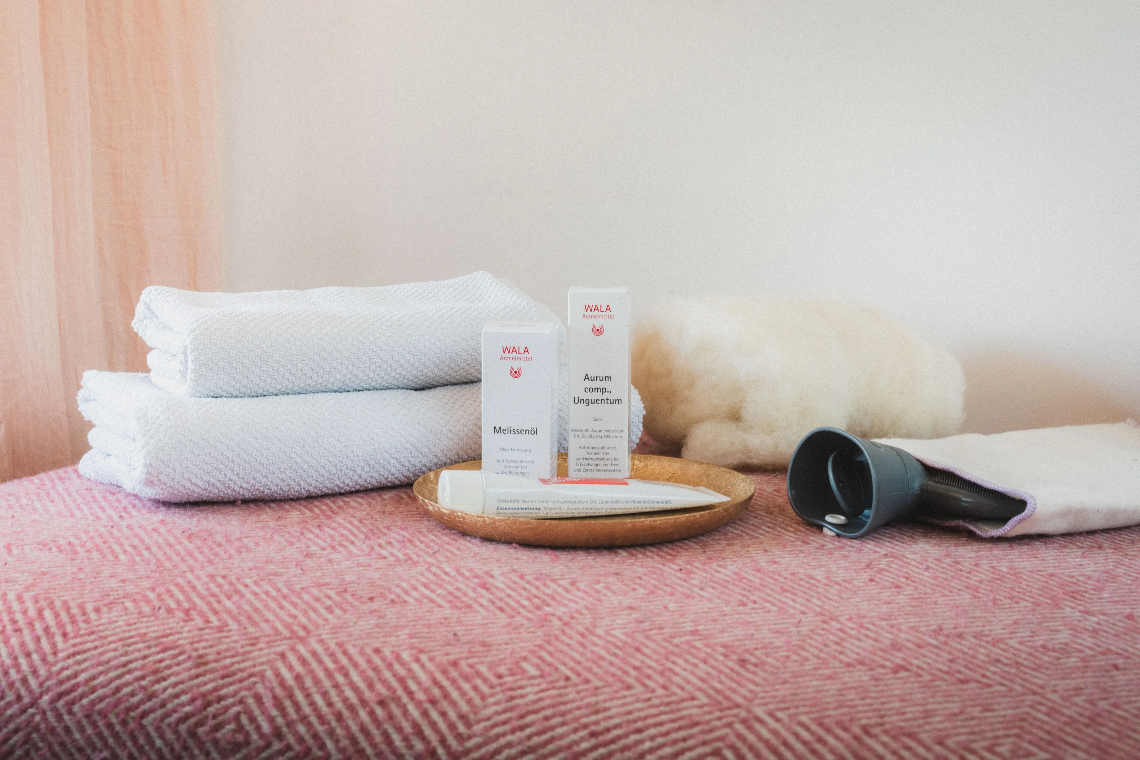 Gefaltete weiße Handtücher, zwei WALA Arzneimittelboxen und eine Tube auf einem goldenen Teller, neben einem blauen Handgerät und einem wolligen Kissen auf einem rosafarbenen Musterstoff.